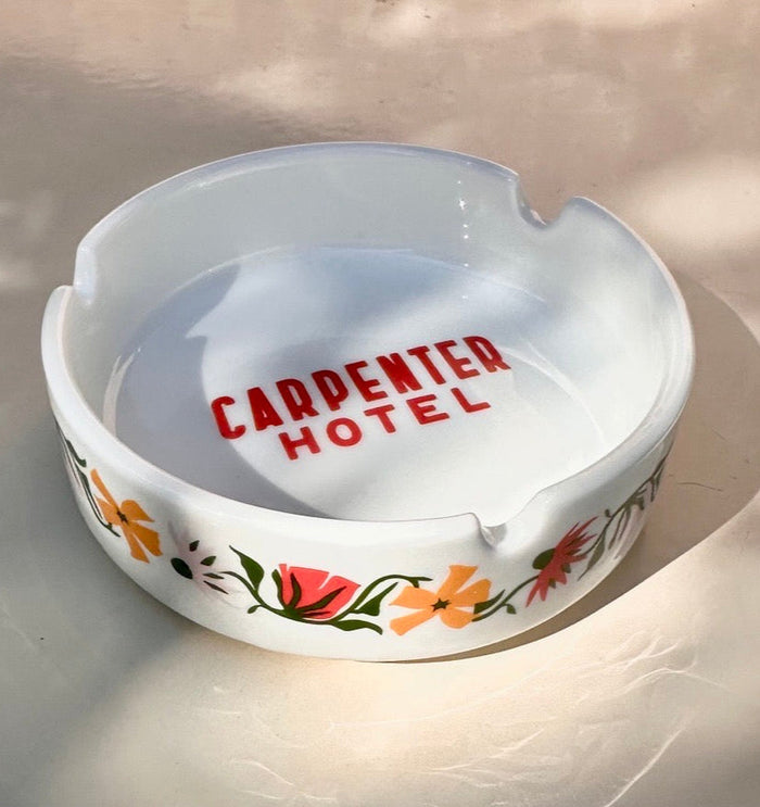 White ceramic ashtray with floral designs on outside and 'CARPENTER HOTEL' text in center bottom sitting on a light background