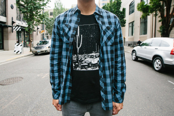 Man wearing black Desert Cruiser tee with white desert scene and jeep, layered under a blue plaid shirt.