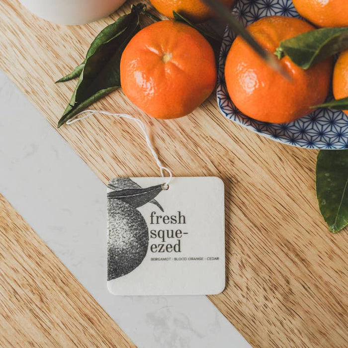 White square air freshener tag with orange graphic beside fresh oranges on a wooden board