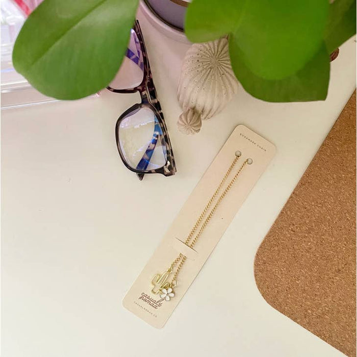 Saguaro Cactus Chain Bookmark shown on a white desk beside a plant, seashells, and reading glasses.