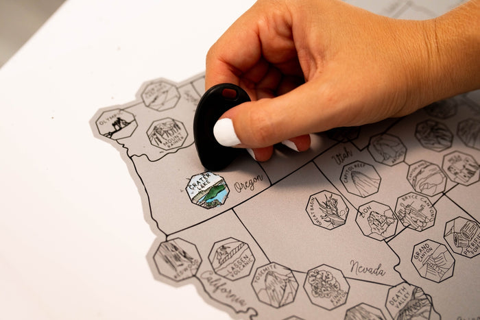 Close-up of a hand using a black scratch tool on a National Parks scratch-off map over Oregon