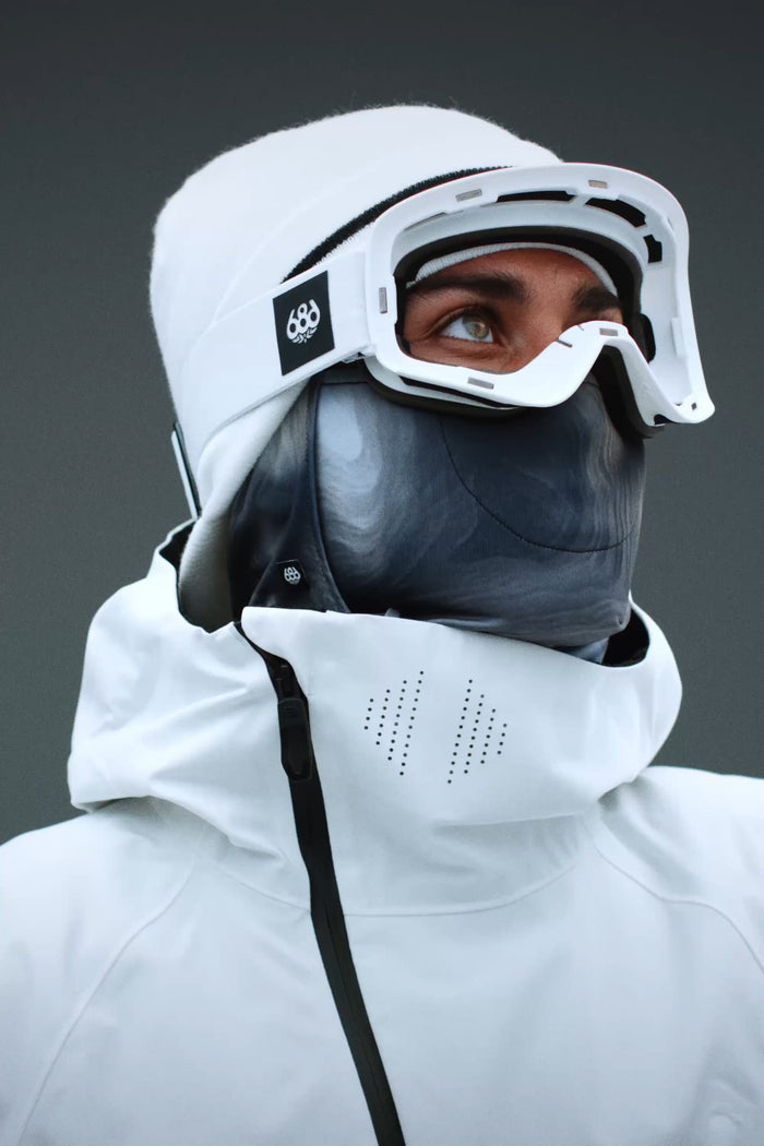 A person wearing a white winter jacket, gray face mask, and 686 SMARTY® Magnetic Goggle by 686 looks upward against a plain gray background.