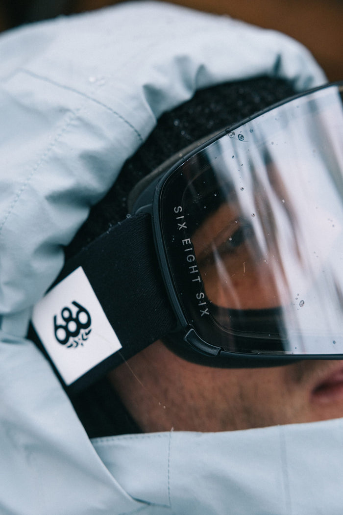 A close-up of a person wearing a blue hooded jacket, black 686 SMARTY® Magnetic Goggle with SIX EIGHT SIX on the strap, and a beanie. The white 686 logo patch is visible on the goggle strap.