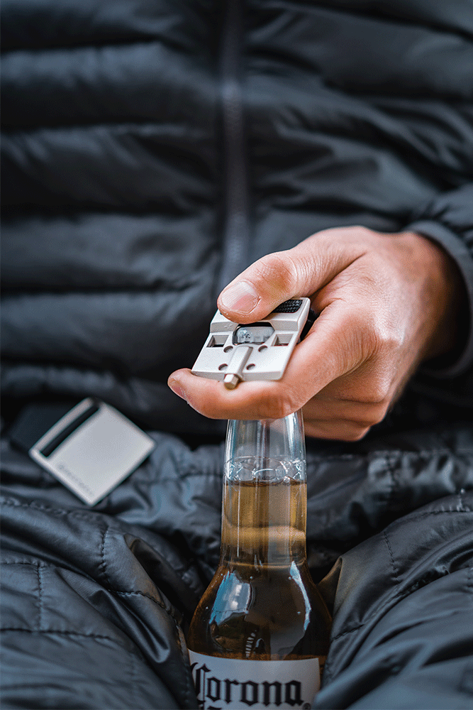 A hand uses the 686 Westwell Clicker Stretch Belt, with a built-in bottle opener, to open a Corona beer bottle against a background of a dark quilted jacket.