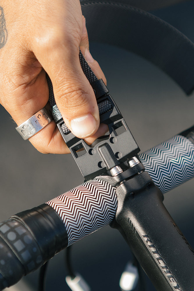 A hand secures the 686 Westwell Clicker Stretch Belt by 686 onto a bicycle handlebar wrapped in patterned tape, fastening it by pressing the buckle with their thumb.