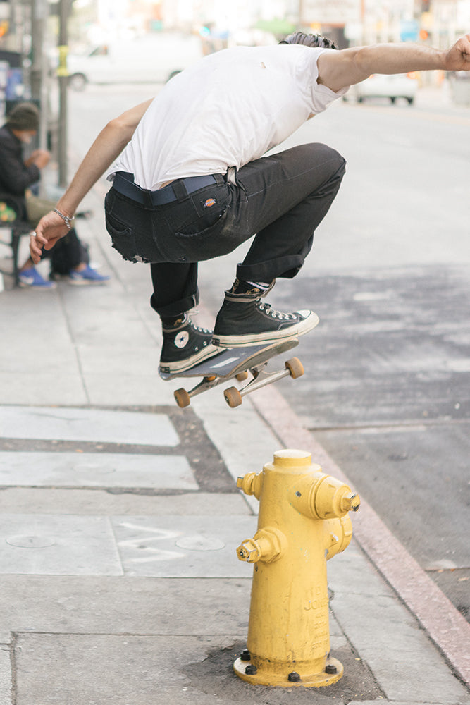 A skateboarder wearing the 686 Westwell Clicker Stretch Belt jumps over a yellow fire hydrant on a city sidewalk, captured mid-air with knees bent and arms out for balance.