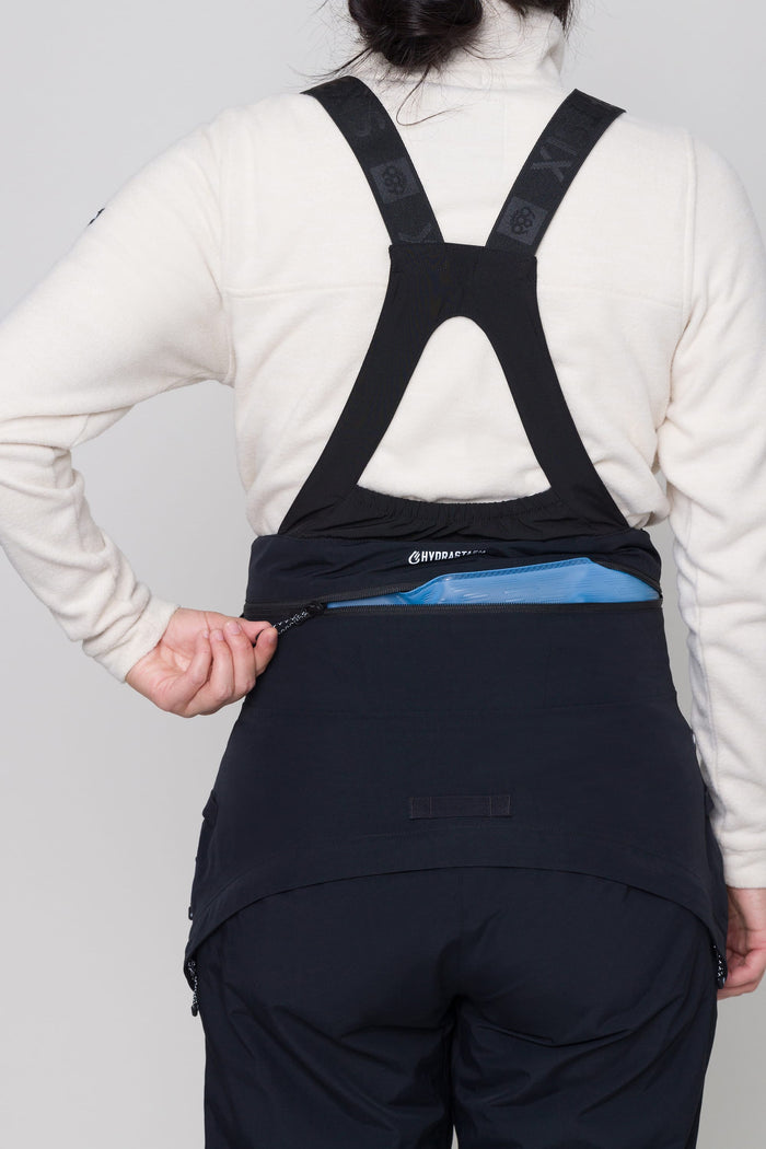 A person wearing a cream jacket and black overalls unzips the blue Hydrastash zipper of the 686 Womens Complete Bladder System by 686 at the lower back of their pants, seen clearly from behind with prominent zipper and strap details.