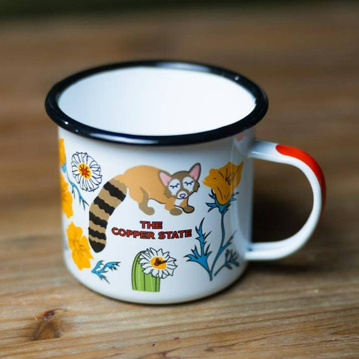 Enamel mug with a dark rim and orange handle adorned with a cat, cacti, and the words The Copper State.