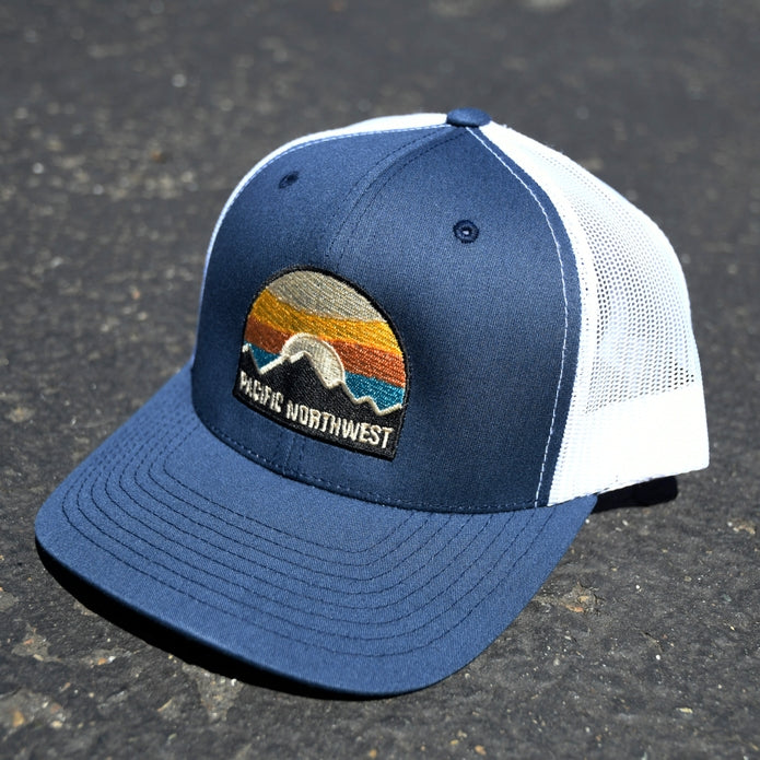 Navy blue curved brim trucker hat with white mesh back and a Pacific Northwest sunset patch.