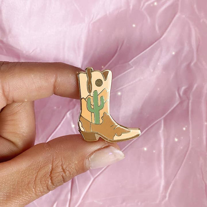 Desert cowgirl boot enamel pin with a green cactus against a pink background.
