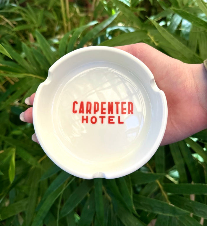 Hand holding a white ashtray with 'Carpenter Hotel' text against a green leafy background