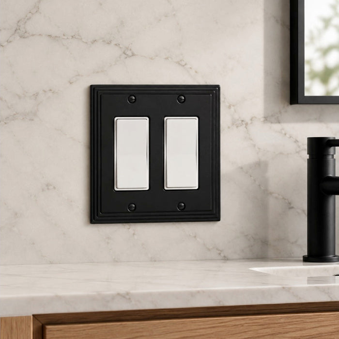 A black double light switch plate cover mounted on a white marble bathroom wall above a marble countertop with a black faucet.