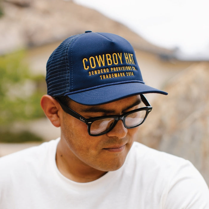 Navy blue trucker hat with yellow embroidery reading COWBOY HAT on the front