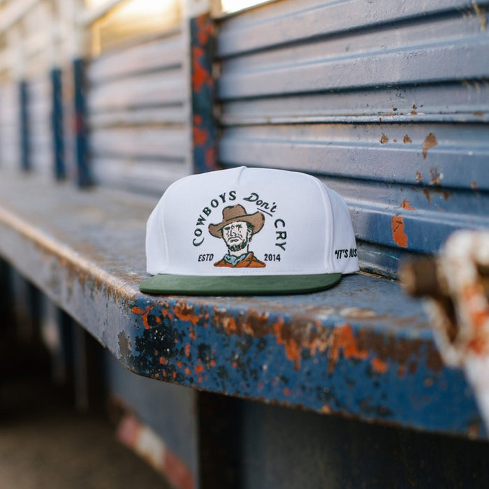 White cap with green brim on a weathered blue metal surface with Cowboys Don't Cry embroidery on the front.
