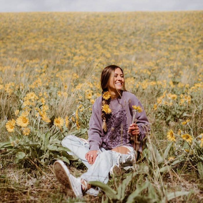 Woman wearing Flower Meadow Crewneck Pullover sits in a field of yellow wildflowers, holding a flower.