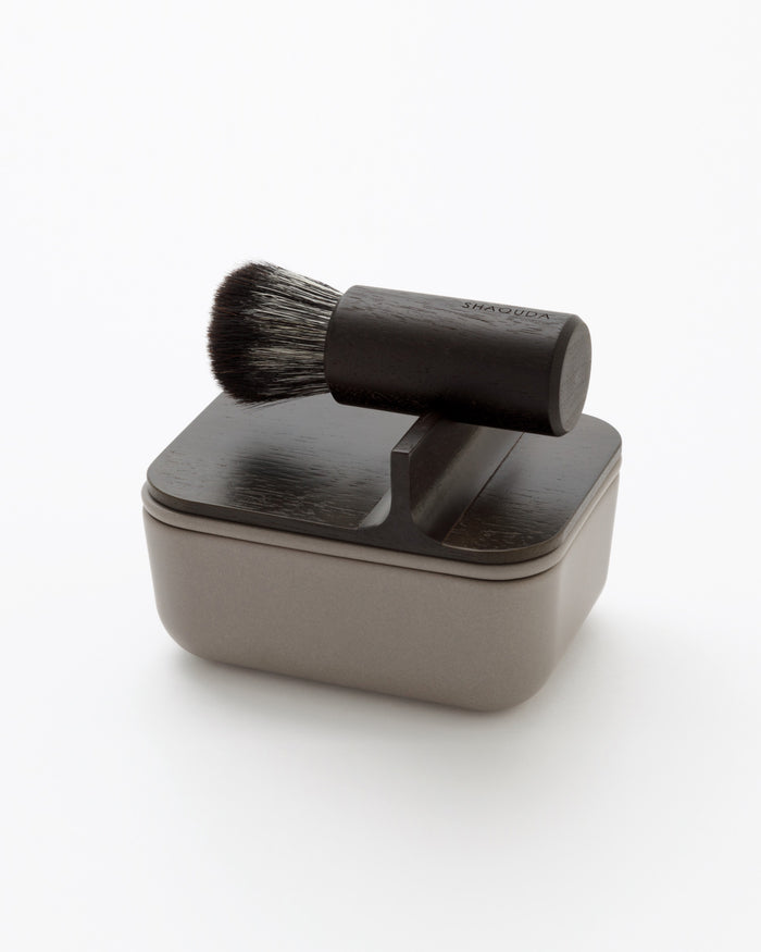 Angled image of hard Jiva face cleansing and shaving series with porcelain bowl and walnut, boar bristle, and goat hair brush by Shaquda against white-gray background.