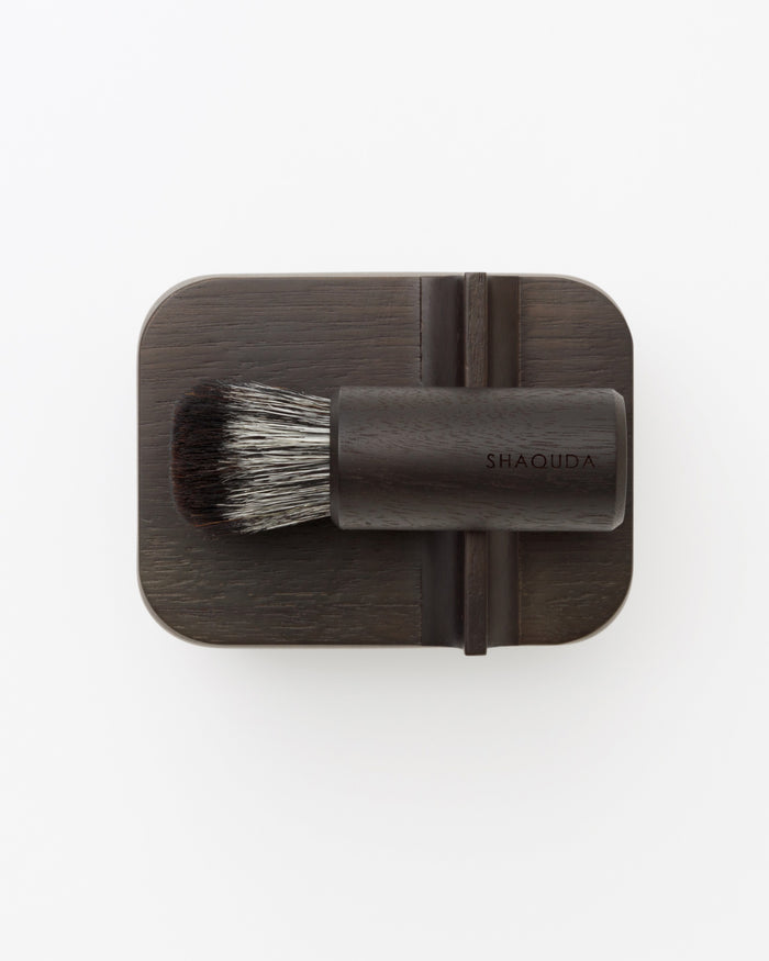 Bird's-eye-view of hard Jiva face cleansing and shaving series with walnut, boar bristle, and goat hair brush on top of porcelain bowl by Shaquda against white-gray background.