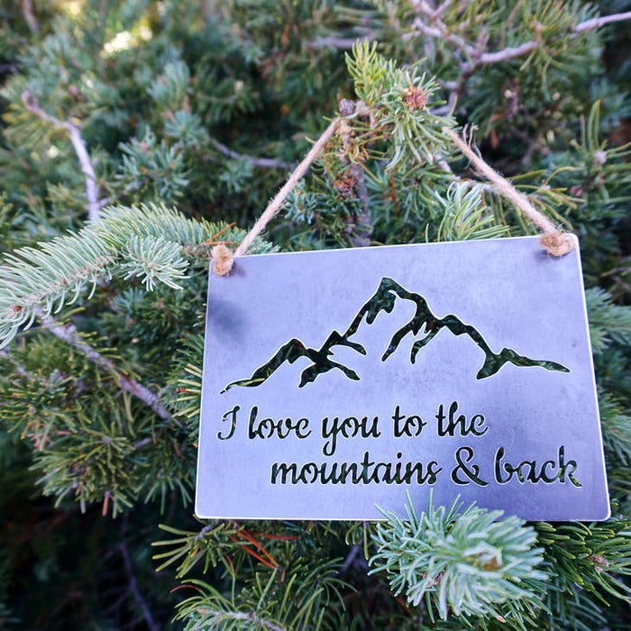 Metal sign with mountain design and the text I love you to the mountains & back, hung on twine among evergreen branches.