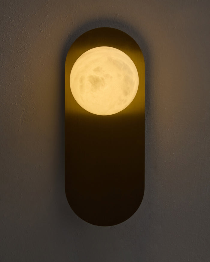 Milan wall sconce lit on a textured wall, featuring a brushed brass vertical backplate and a glowing round white glass globe that casts a warm ambient light.