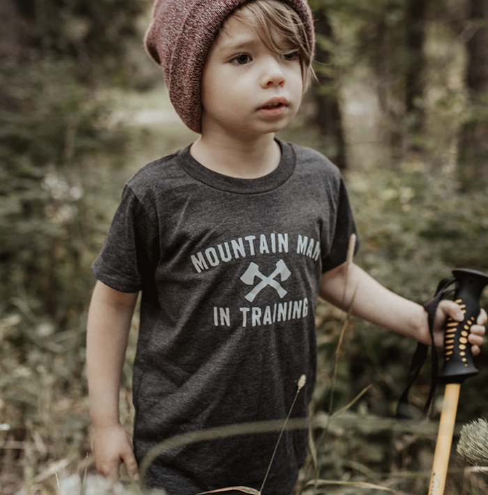 Toddler wearing a dark gray Mountain Man in Training tee with crossed axes outdoors
