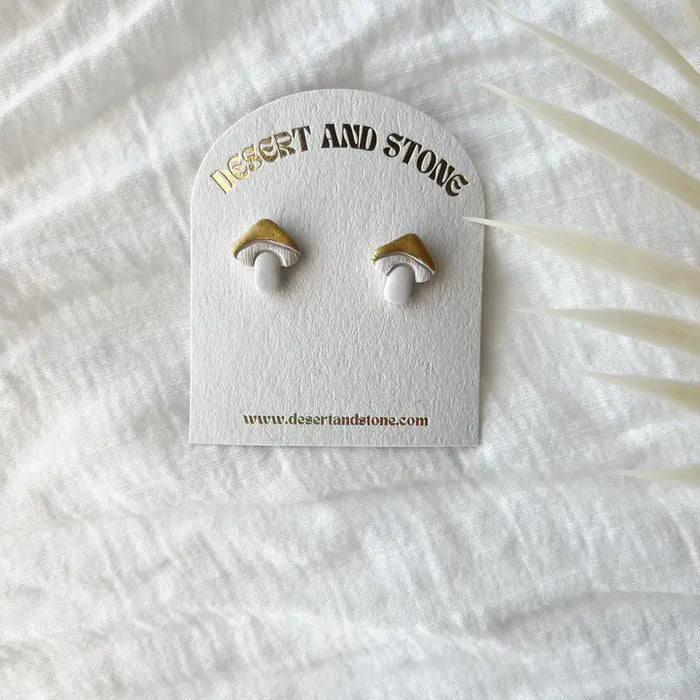 Two mushroom shaped stud earrings in gold and white on a Desert and Stone branded card