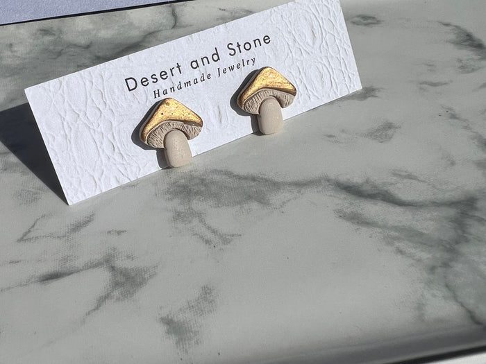 Gold mushroom stud earrings with white stems mounted on a textured Desert and Stone display card