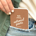 Brown national parks journal held in a denim pocket by a hand with white nails.