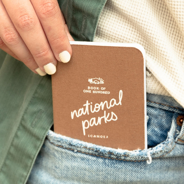 Brown national parks journal held in a denim pocket by a hand with white nails.