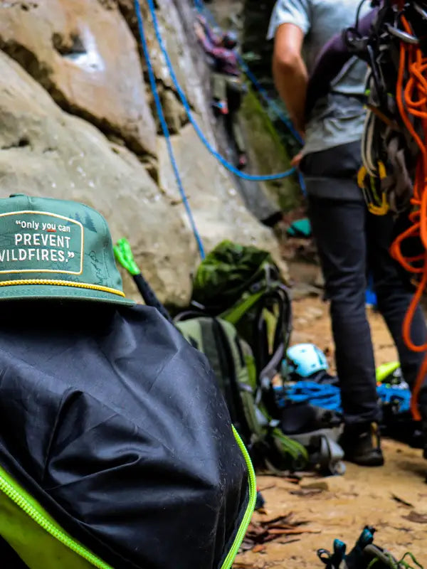Green climbing hat with a patch saying only you can prevent wildfires among ropes and gear in a rocky outdoor setting.