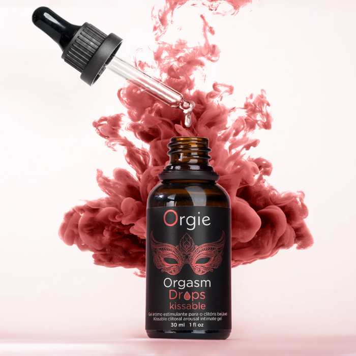 A 30ml amber bottle of Orgie Orgasm Drops centered against a light pink background. A glass dropper is angled above the open bottle with a single clear drop falling into it. Directly behind the bottle, a vibrant, billowy cloud of reddish-pink ink or smoke explodes outward, creating a dynamic and sensual visual effect. The label identifies the product as a "Kissable clitoral arousal intimate gel".