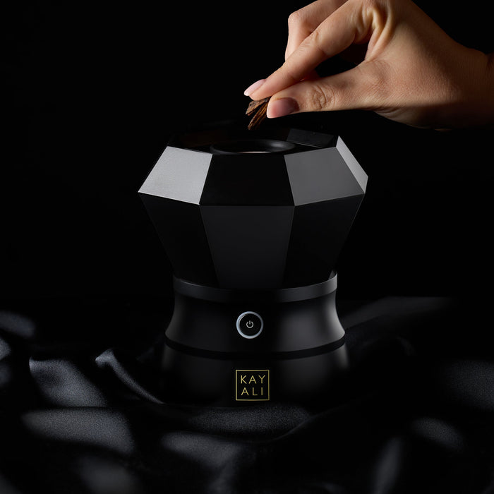 Person placing bahkoor into a black and white geometric-shaped container with 'KAYALI' branding.