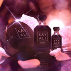 Two bottles of Kayali oudgasm rose oud perfume on a dark surface with blurred lights and flowers in the background.