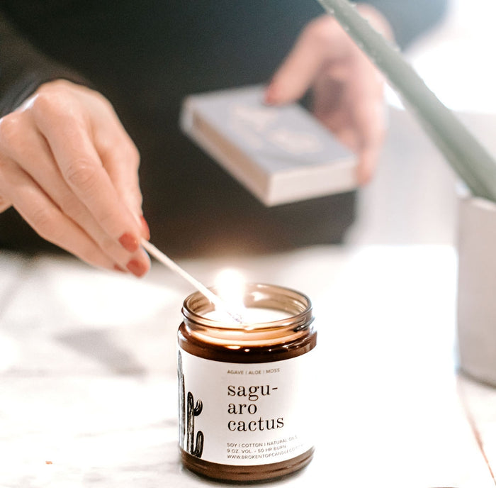 Hands lighting the Saguaro Cactus candle with a match beside a white box