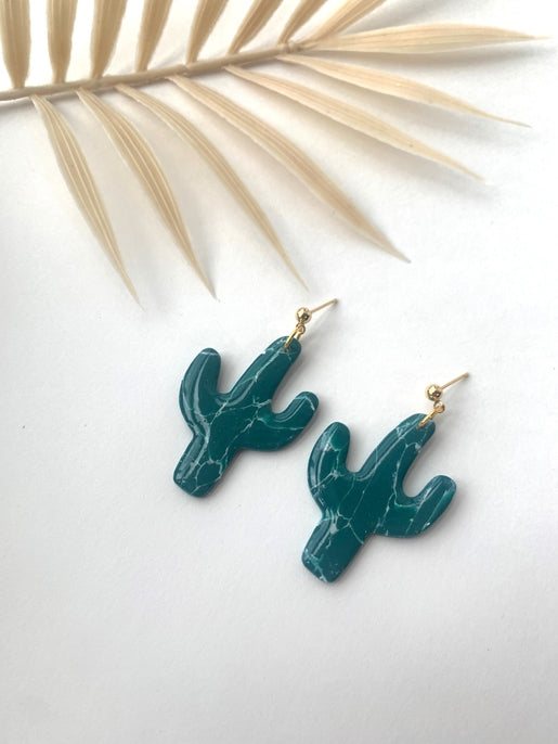 Teal cactus shaped earrings with white veining and gold hooks on white background