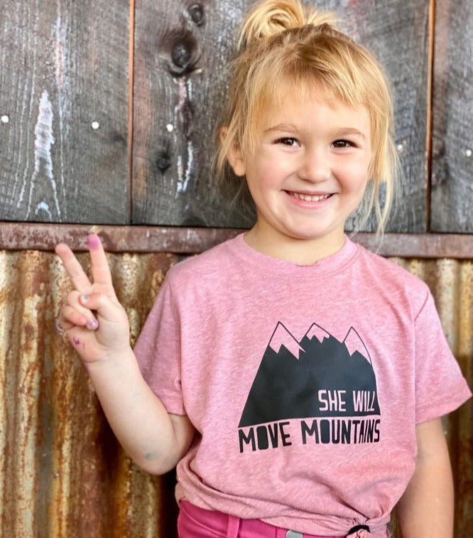 Smiling girl in pink kids tee with mountain graphic and She Will Move Mountains text.