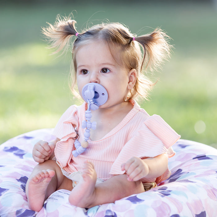 A baby with light brown hair in pigtails sits outdoors on a floral-patterned cushion, wearing a pink top and using the Sili Soother Pacifier (2 Pack) with a beaded clip.