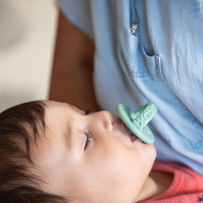 A baby sleeps peacefully with closed eyes, sucking on a Sili Soother Pacifier (2 Pack) in green, while resting against an adult in a light blue shirt.