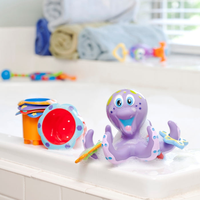 The Splish Splash Stacking Cups - 5 pack sits in a bubbly bathtub next to a purple octopus bath toy with colorful rings, making bath time fun and engaging for baby development. Rolled towels and extra bath toys are by the window.