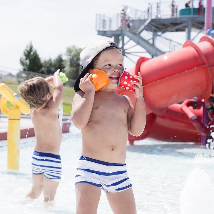 Two young children in swimsuits play in a splash pad. The child in front wears a backwards cap and holds Splish Splash Stacking Cups - 5 pack, with slides and water features seen in the background.