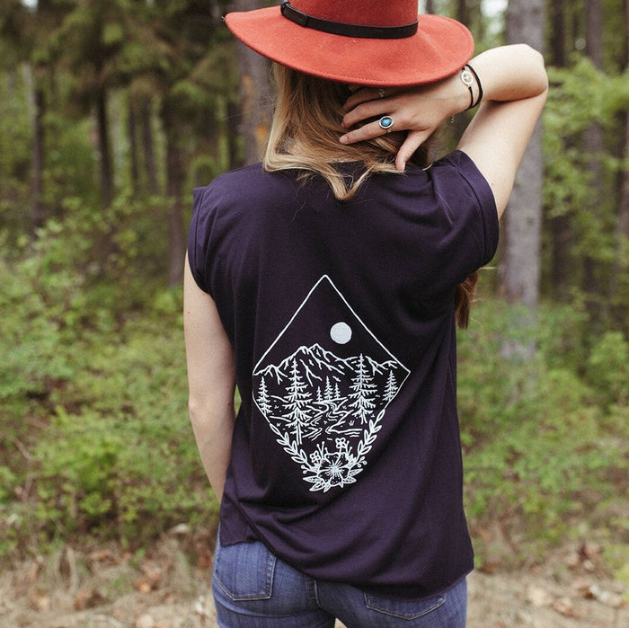 Back view of a person in a dark purple Take Me to the Wildflowers tee with a white mountain forest graphic in a wooded setting with a red hat.