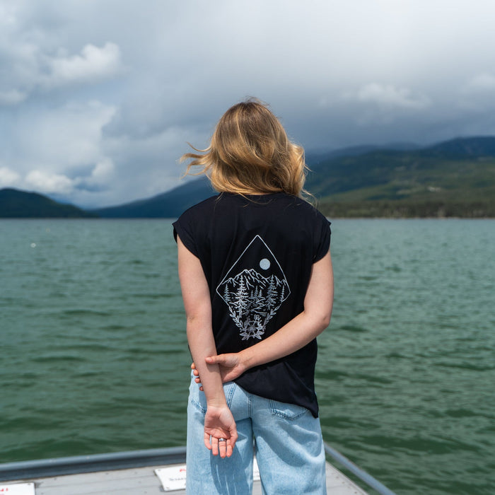 Person with back to camera wearing black Take Me to the Wildflowers rolled sleeve tee by a lake with mountains in the background
