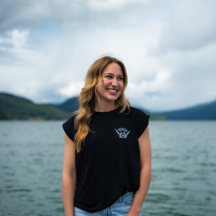 Smiling woman in black Take Me to the Wildflowers tee with a white emblem on the chest by a lake and distant mountains