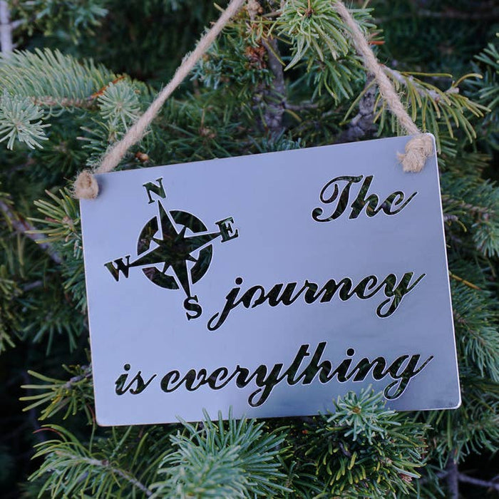 Small gray metal sign with a compass design and the words The journey is everything, hung from rope among evergreens.