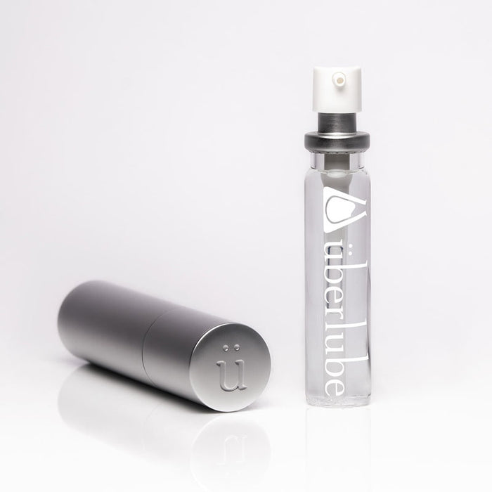 A clear "überlube" bottle with a white pump top stands upright next to a closed silver cylindrical travel case featuring the "ü" logo on its end, both products available from Worldcondoms.