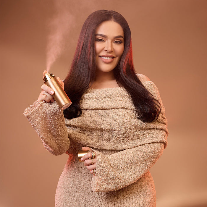 Mona Kattan holding a gold hair spray bottle against a brown background