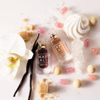 Two perfume bottles labeled 'KAYALI' surrounded by vanilla beans, flowers, and candies on a white background.