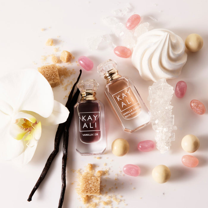 Two perfume bottles labeled 'KAYALI' surrounded by vanilla beans, flowers, and candies on a white background.