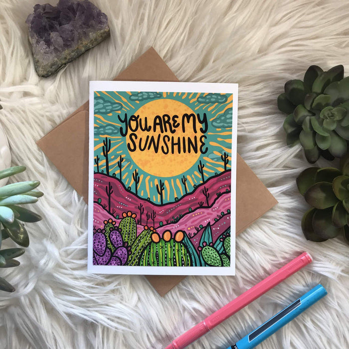 You are my sunshine greeting card with a bright yellow sun, turquoise sky and purple desert hills with cacti.